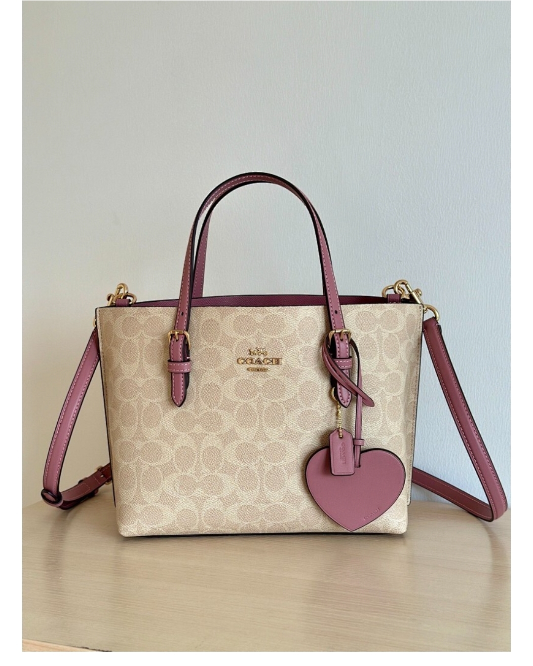 Beige signature print tote bag with pink handles, gold hardware, detachable strap, and heart-shaped charm detail.
